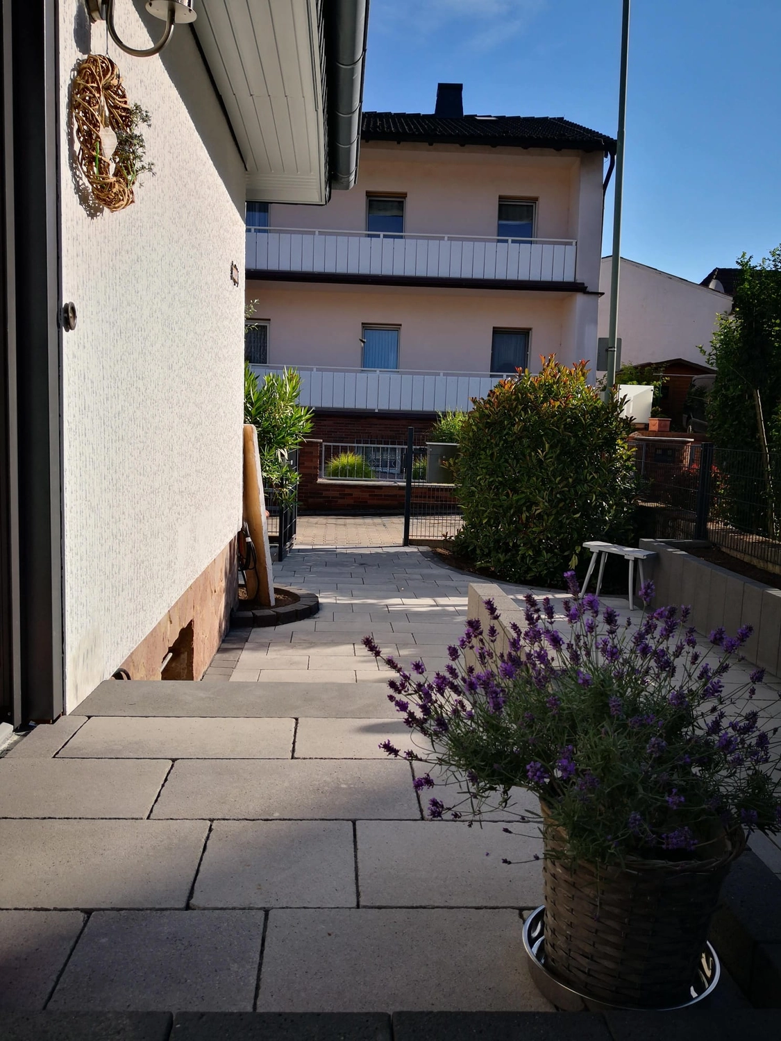 Zois Gartengestaltung – Dieser ruhige Außenbereich verfügt über eine gepflasterte Terrasse, die mit einem Lavendeltopf und einem Kranz an der Mauer geschmückt ist.