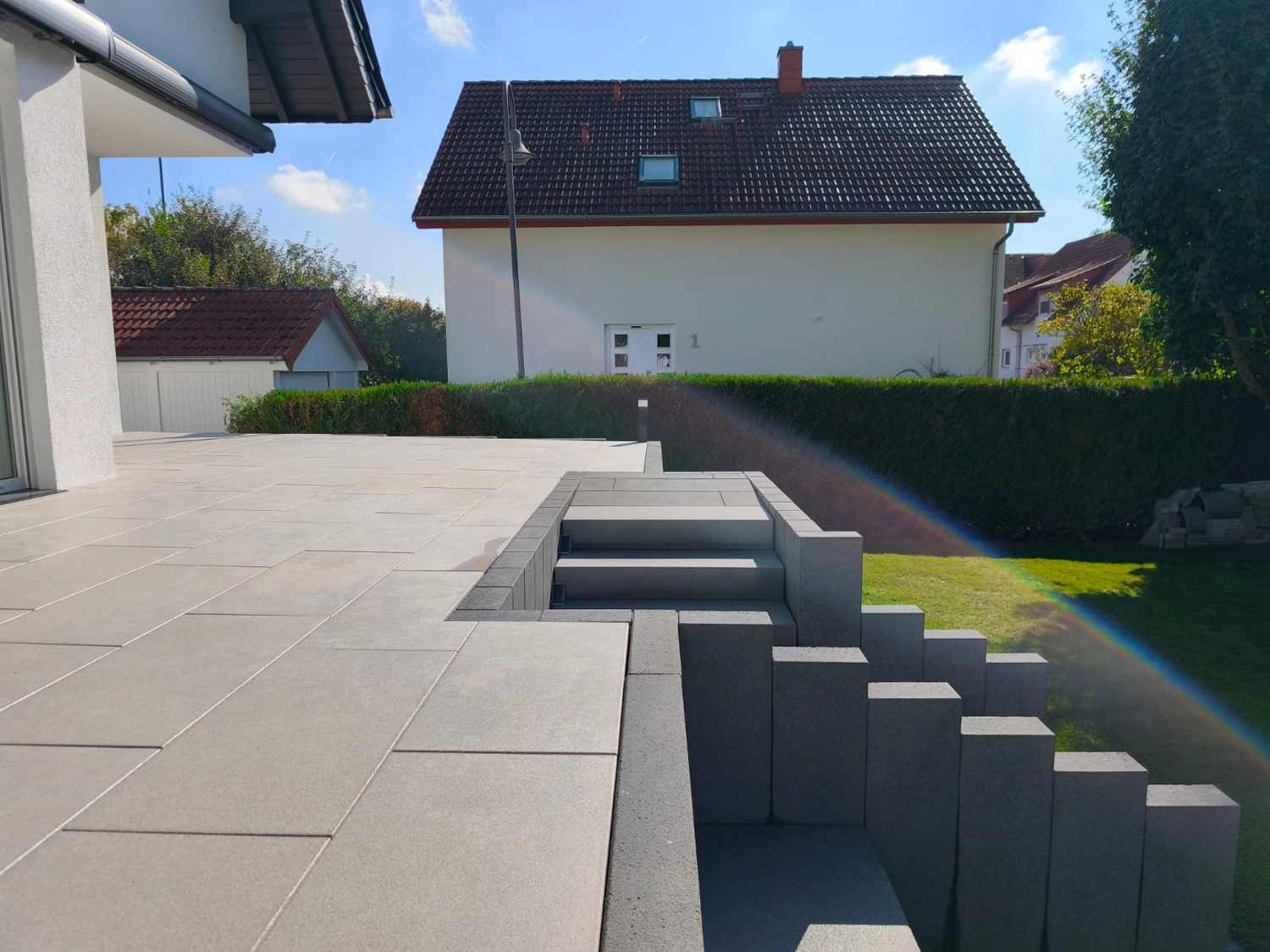 Zois Gartengestaltung – Diese moderne Betonterrasse ist mit großformatigen Pflastersteinen und gestuften, L-förmigen Stützmauern versehen, die zum Rasen hinabführen.